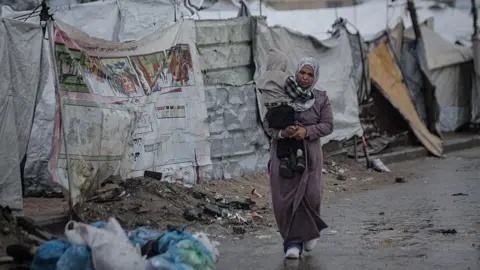 Anadolu via Getty Images A displaced Palestinian woman carries a child as she walks past makeshift tents. There is rubbish on the floor and puddles as she walks. 