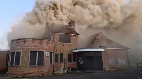 An empty red brick pub with blocked up windows, with smoke coming out of it. There is graffiti on the walls. The smoke is brown and thick. One part of the building is round, with different sections to the building. 