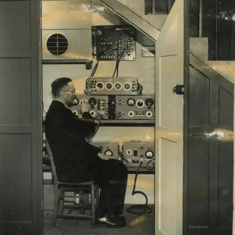 St Martin-in-the-Fields Black and white image of a man sitting in a cupboard beneath some stairs moving dials on boxes of radio equipment which are on shelves