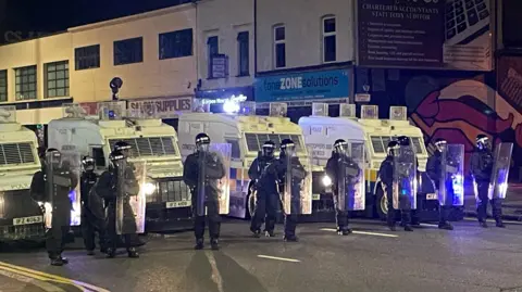BBC A line of police officers in riot gear, with shields, stand in front of a row of armoured police Land Rovers