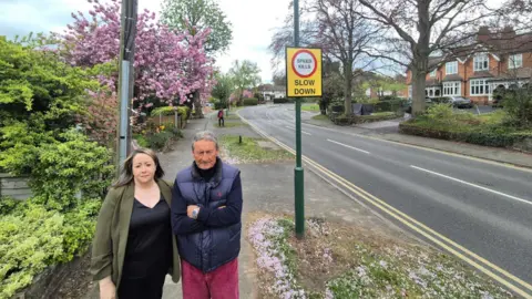 A man and woman stand on the pavement to the side of a road in a residential area. There is a sign by the road saying "Speed kills, slow down."