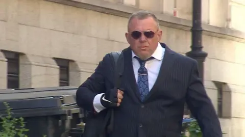Mark Killick walks towards Bristol Crown Court. He is wearing a dark grey suit and white shirt with a dark blue tie. He is clean shaven and wearing sunglasses and has short, cropped hair