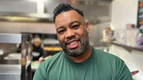 A man with black and grey hair and a beard is wearing a green t-shirt. He is stood in a kitchen.