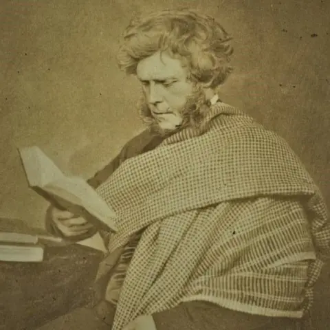 Getty Images An albumen print of Hugh Millar, photographed by J.G. Tunny between 1855 and 1857. He seated at a desk wearing a checked shawl draped over his shoulders over a dark suit and looking down to an open book held in his right hand. He has wavy hair covering his ears and thick sideburns. 