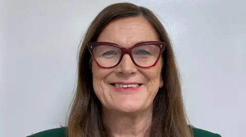 Joanne McMullan A woman with long brown hair is looking into the camera. She is smiling. She is wearing red-rimmed glasses and a green jacket is visible at the top of her shoulders. She is standing in front of a plain white wall, it appears to be indoors. 