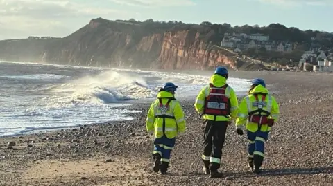 BBC Three emergency service workers dressed in hi-visibility clothing and blue hard hats walking on a stone beach. They are wearing fluorescent clothing. A clifftop is seen in the background. There are large waves breaking onto the shore.