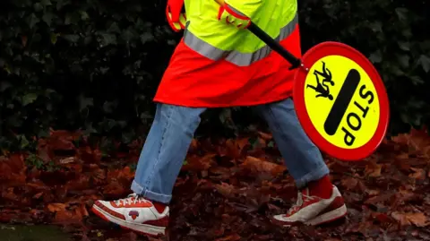 A general view of the feet of a school crossing patrol man, known as a Lollipop Man, carrying a sign that reads: STOP and has the image of two children underneath.