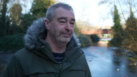 Steve Sheldon is wearing a green coat with a furry hood and is standing next to the underpass