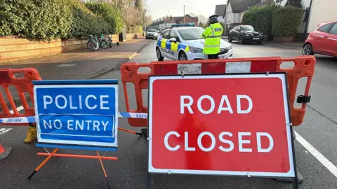Richard Knights/BBC A blue "Police No Entry" sign and a red "Road Closed" sig in front of barriers put across a road. A police officer with his back to the camera stands in front of a marked police car. 