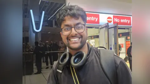 Man in his 20s with dark hair and neat beard wears a black t-shirt and backpack with headphones around his neck. Wearing large, dark-framed glasses, he is standing by the arrivals section at Manchester Airport.