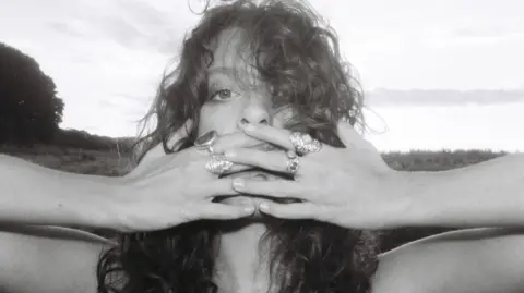 Heidi Curtis Heidi Curtis has long curly hair and she is covering her mouth with both hands. She has four rings, two on each hand. The picture is in black and white and a field can be seen behind her.
