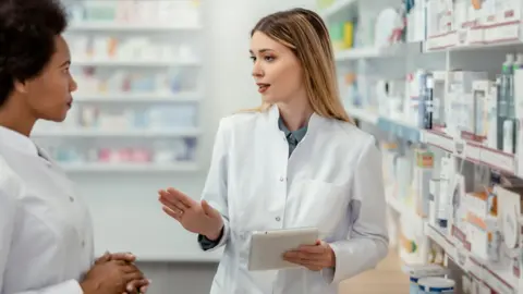 Woman in white lab coat with blonde hair, chats to woman in white lab coat with brown hair, in pharmacy surrounded by products.