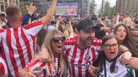 Sunderland fans at the fan zone in the city. Two women and a man - wearing Sunderland colours - are shouting in celebration. Behind them are the backs of fans facing a giant screen. Most have hands raised and many are holding mobile phones aloft.