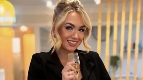 A smiling Ashleigh Watson pictured in a soft focus shot holding a flute of champagne. She has blonde hair and is wearing a black blouse and matching skirt.