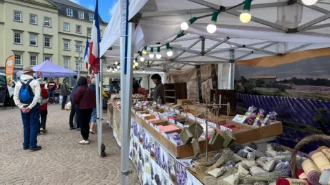 Market stands side by side. There are soaps and clothes on the stalls. There are people walking around. 