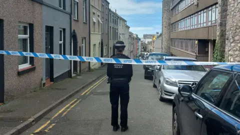 A police officer facing away from the camera in a closed terraced street in Caernarfon, near vehicles, and with blue police tape across the road
