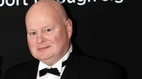Nigel and his wife Penny pose for a picture at an event. A bald man, he wears a black tuxedo jacket and bowtie over his white dress shirt with black buttons. Penny has short dark hair and smiles while wearing a gold evening dress.