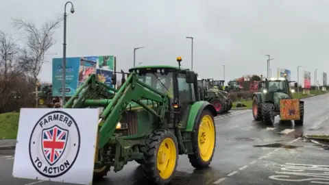 Tractors are in a queue in a car park at the Cornwall Services. The first in line has started the procession and it has a large sign reading FARMERS TO ACTION with a shield formed of the Union Jack flag tied to its bucket.