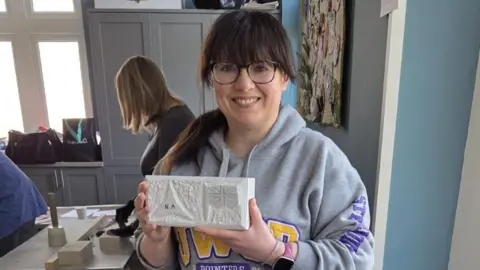 Rob MacDonald Nicola Andrews is smiling as she holds a white rectangle cobble she made for the monument's base. She has glasses and a grey hoodie. Her straight brown hair is tied in a ponytail and she has a fringe. The design on the cobble includes a triangular shape and the union jack flag.