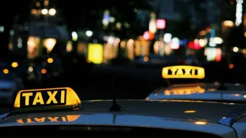 The top of two vehicles which have yellow and black glowing light signs on them, reading 'taxi'. City streets can be seen in the background, quite blurred. 