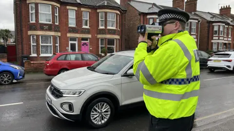 PC Daniel Brock stands in his high vis and police uniform on the side of the road looking down the barrel of the police speed gun. Cars pass on both sides of the road showing how busy it is. 