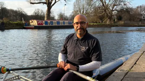 Ben Schofield/BBC Omar sitting in a single scull rowing boat on the side of a river. He is smiling slightly, while holding an oar in each hand. It is a fine, bright day. He is wearing a dark blue, collared t-shirt, with all three buttons done up, though his arms are covered by a white top worn underneath the t-shirt. He is wearing dark blue jogging bottoms and the tops of his knees are just in shot. His boat is against a pontoon edge, which is to the right of frame. Behind Omar is the far river bank, with a narrow boat moored up, a grassy bank, trees and a building. The river looks calm and it is a bright day.