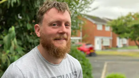 BBC A man with a ginger beard and brown hair, wearing a grey t-shirt, standing in front of a tree with a red car and houses in the background.