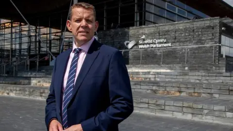 PA Media Plaid Cymru's leader Rhun ap Iorwerth standing in front of the Senedd building in Cardiff Bay. He is wearing a navy suit and a pink shirt, with a striped tie.  It is a sunny day.