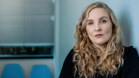 BBC Tamsin McNally looks into the camera. She has long, wavy blonde hair and is wearing a plain black top with blue chairs visible over her right shoulder. 