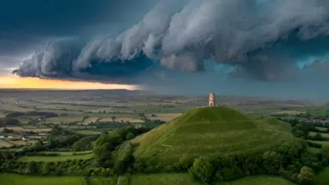 Glastonbury Tor in the middle is surrounded by green fields and trees and above there is a stunning grey storm cloud in a dark sky with a glimmer of sunlight on the horizon
