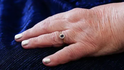 Close up of Pamela's hand with a gold ring on her fourth finger. Her nails are painted silver. 