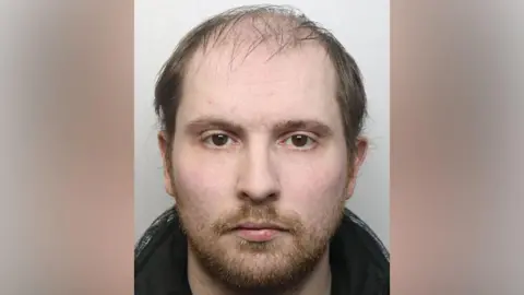 A police profile photo of a man with a auburn beard wearing a black jacket.