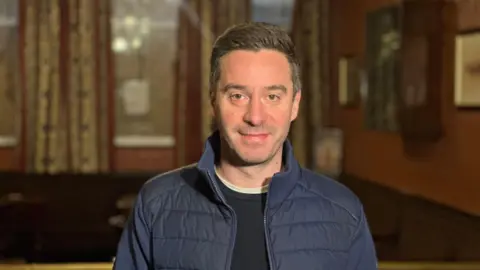 James Graham has brown hair and brown eyes. He is wearing a dark blue, lightweight puffer jacket. He's wearing a black t-shirt with a white one underneath. He's standing in a pub smiling. 