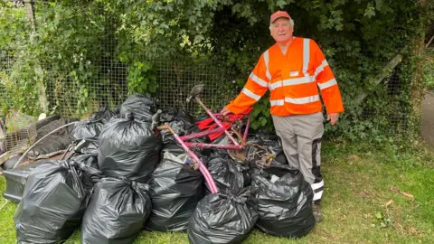 John Devine/BBC A man is standing on a grass verge next to numerous black rubbish sacks and part of an old pink bicycle. He is wearing an orange jacket, an orange cap and beige trousers.