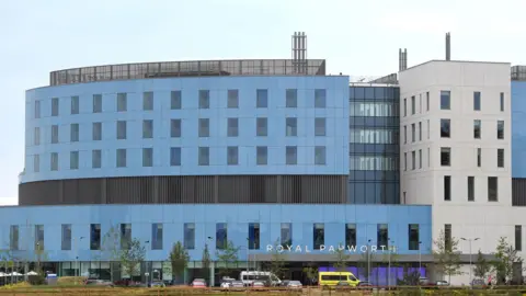 PA Media The front facade of Royal Papworth Hospital. The picture is taken from at least 200 metres away, and it shows a large part of the multi-storey building. It is blue and white with a glass walkway separating two sections of the building. The building is curved and is six stories high. You can see ambulance transport vehicles parked outside the front.