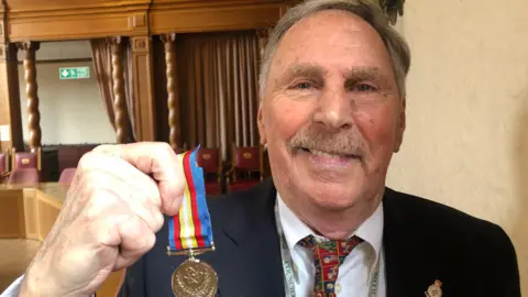 Alan Dowson, who has short grey hair and a moustache, smiling into camera holding up Nuclear Test Medal. He is wearing a navy suit, white shirt and red-patterned tie. 