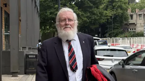 An old man with white grey hair, beard and moustache wearing glasses and a suit with a stripy tie is walking down a road in a city centre, next to a row of cars.