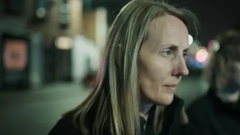 Blast Films Crime scene coordinator, Jo Ward, is standing in a street. We see only the right side of her face. She has shoulder length blonde hair and is wearing a black top. She is listening attentively to someone who is not in the shot. In the background, you can see properties. it is evening time and the night sky behind Jo is dark.