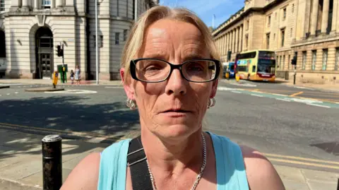 A woman wearing a turquoise vest and a gold chain around her neck stands on a pavement in front of a road junction facing a court building. She has blonde hair tied back and black-rimmed glasses. Behind her, grand stone buildings can be seen on the opposite side of the road, while two buses are waiting at the junction.