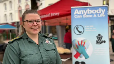 Ms Campbell is wearing a green ambulance shirt. She has long brown hair which is tied up. She is wearing glasses. The ambulance worker is stood in front of a blue sign which says anybody can save a life.