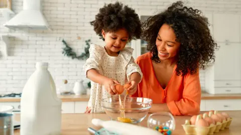 Getty Images A child and her mother sat on a kitchen table, the child wears a white polka dot dress and the mother wears a bright orange blouse, the child is cracking an egg into a bowl.