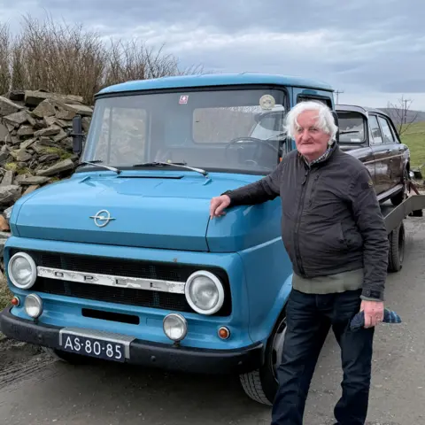 A man with white hair leans on a vintage blue Opel truck. It has a mulberry-coloured car on the back of it. 