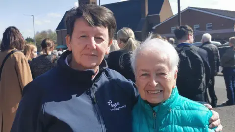 Two women are standing together. One has short brown hair and is wearing a navy hoodie saying age Ni. The other has short white hair and is wearing a green jacket. People are standing in the background. A church is across the street. 