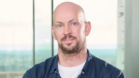 James Watt has a bald head and a beard. He is wearing a blue shirt over a white T-shirt and is sitting in front of a window