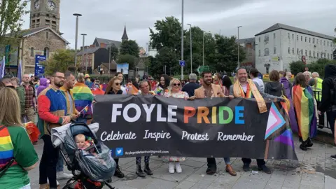 Un grup de oameni ținând un banner Foyle Pride în timp ce stau în fața gării Derry, cu o mulțime mare în spatele lor. O serie de clădiri pot fi văzute în fundal, în timp ce un bărbat și un copil într-un cărucior stau în dreapta lor