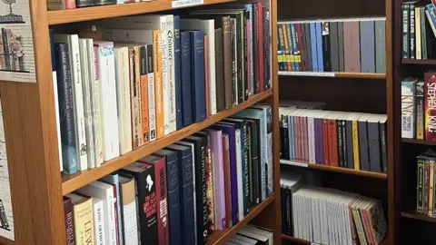 In the foreground is a large wooden bookcase which has several rows of books on it. In the background there are rows of bookshelves stocked with books.