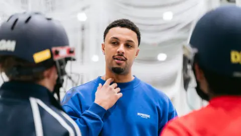 Bedford School Emilio Gay, wearing a blue jumper, talking to two children, who have their back to the camera and are wearing cricket helmets. He has one hand to his neck, and he has short hair and a goatee beard. 
