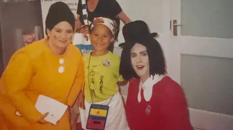 Mali Ann Rees Mali Ann Rees as a child with them Urdd Eisteddfod characters Sali Mali and Jac y Jwc. Sali wears an oversize orange dress and a black wig, and Jac wears a red shirt, black wig and top hat. 