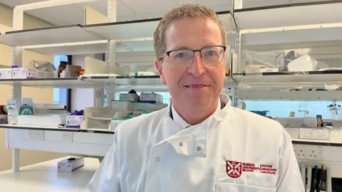 Prof Christopher Scott. He has glasses and short grey-brown hair. He is wearing a white lab coat with a Queen's University Belfast logo on the left breast. In the background there are shelves with various bit of laboratory equipment on them