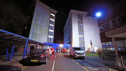 PA Media Two, tall grey and white building towers, face each other, with a stretch of road with hospital drop off parking in front of it. It is night time 